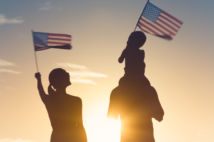A family facing a sunset waving american flags in the air. A child sits on top of his fathers shoulders holding and waving an american flag
