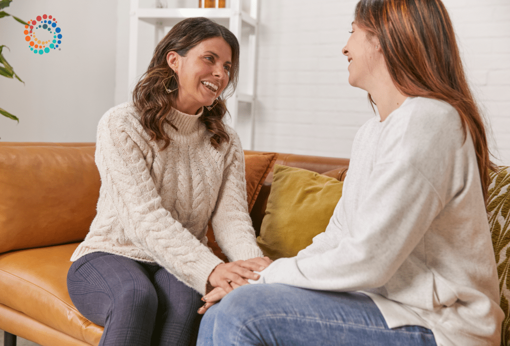 A health coach sitting on a light brown leather couch with her patient holding her hand with a smile on her face.