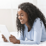 African amercian woman laying on her stomach looking at a tablet smiling and reading what is on the screen