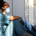 Healthcare professional sitting on the ground in a hallway looking down with a mask on exhibiting the sense of burnout